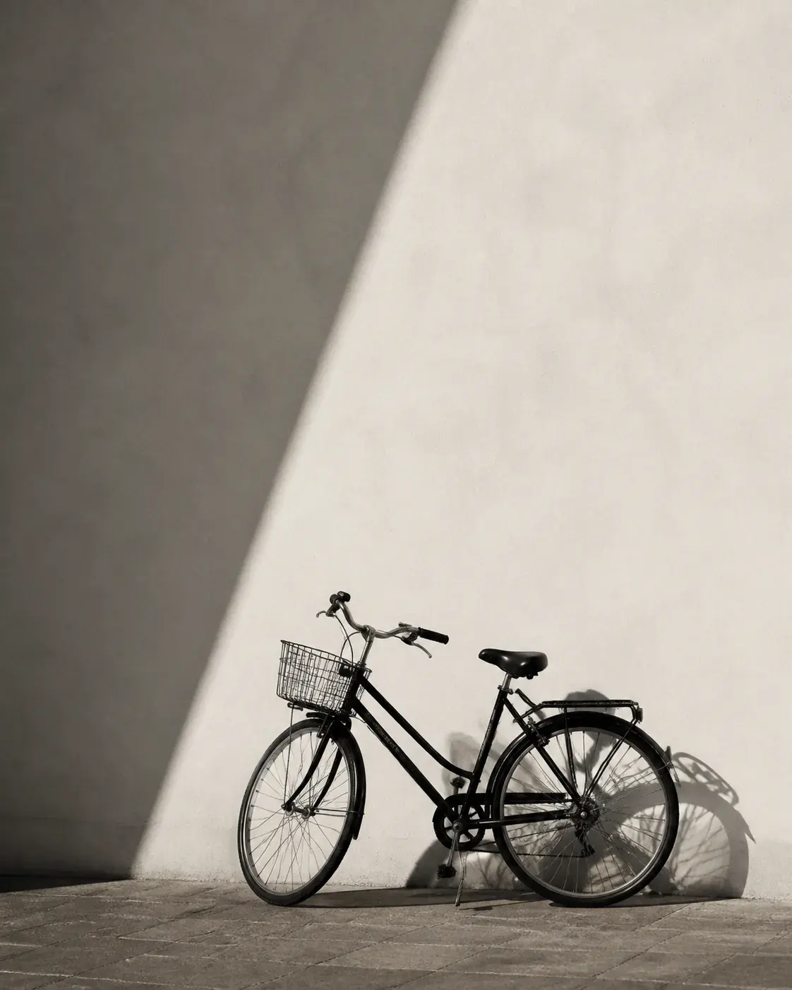 Minimalist monochrome photograph of a black city bicycle against a warm off-white wall with one crisp diagonal shadow, strong