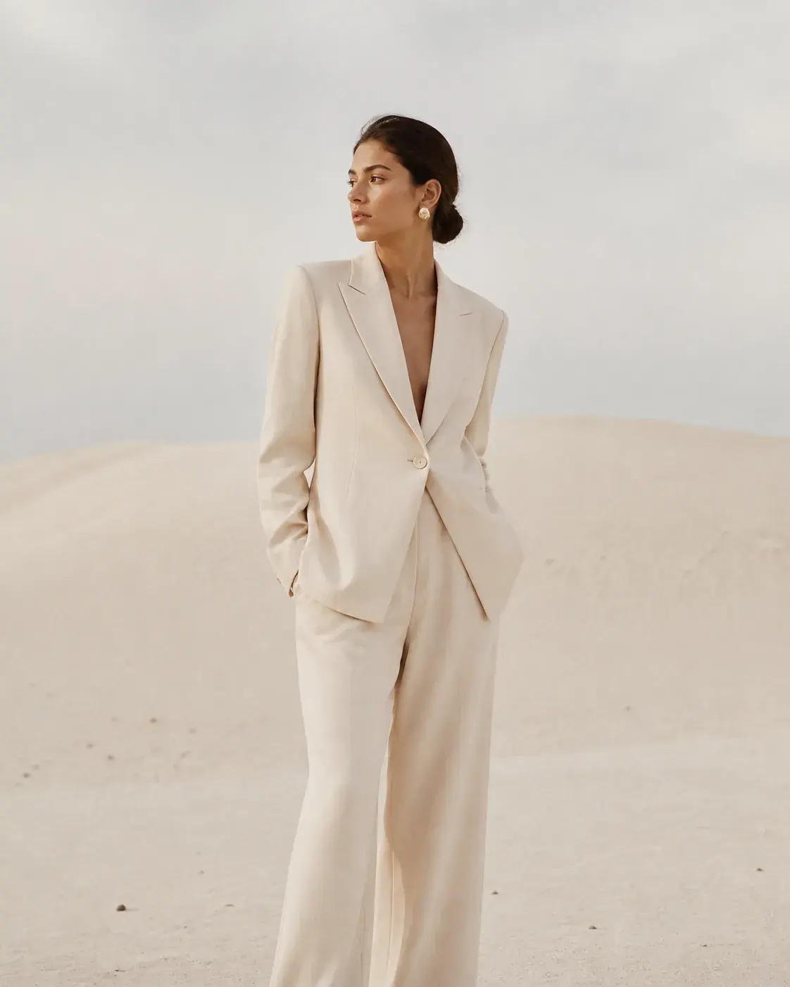 Minimalist editorial fashion photograph of a fictional adult woman in a structured ivory suit standing in a pale sand landsca